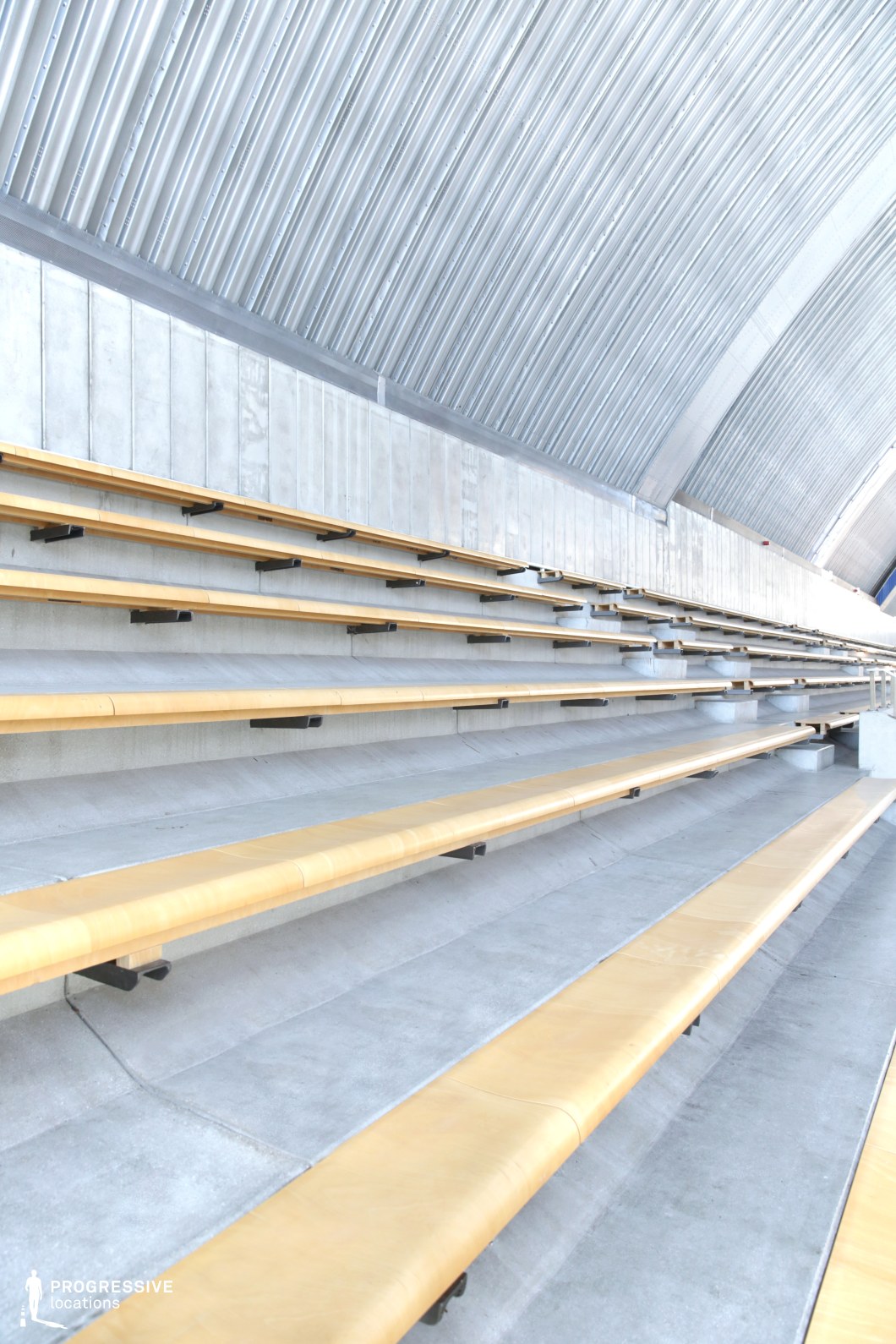 This image captures a section of indoor bleachers defined by clean lines and a highly industrial aesthetic. The seating consists of narrow, light wood benches affixed to a stepped concrete base, with dark metal brackets providing support. Above, a corrugated metallic arch ceiling amplifies the minimalist and futuristic tone of the space, reflecting light uniformly across the structure. The materials—raw concrete, exposed metal, and unembellished wood—create a restrained but modern setting. This location is ideal for productions seeking a stark, geometric visual language or a neutral architectural backdrop that emphasizes symmetry, repetition, and structure.