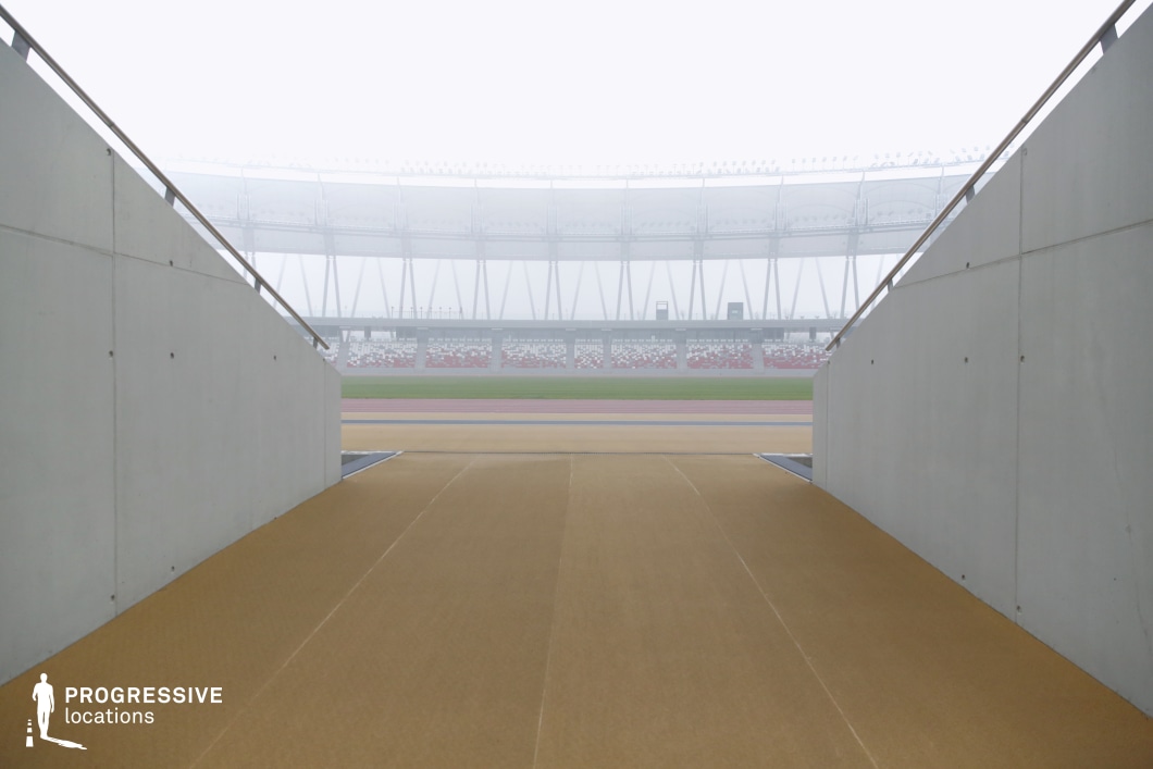 This image captures the tunnel-like entrance to a large-scale athletic stadium, designed specifically for player or performer access. Framed by clean, angled concrete walls and metal railings, the composition leads the eye toward the open field beyond, creating a natural visual funnel that heightens a sense of anticipation and direction. The path is covered in a neutral-toned surface, suitable for heavy foot traffic and equipment transit. In the background, the curved stadium stands and track are partially visible through light atmospheric haze, enhancing the depth and scale of the scene. The red-and-white spectator seats and modern tensile roof structure echo the venue's overall architectural rhythm. This perspective, aligned along the central axis of the ramp, emphasizes geometric balance and clean symmetry. The combination of concrete textures, linear forms, and transitional space makes this an ideal filming location for entrance sequences, sports-related scenes, or settings that require a minimal yet impactful spatial dynamic.