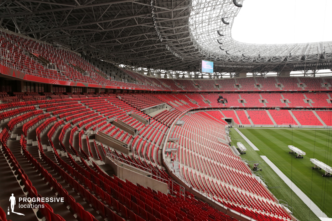 This expansive football stadium interior features sweeping rows of red seating distributed across three distinct tiers, curving uniformly around a well-maintained grass pitch. The camera angle emphasizes the elegant geometry of the bowl-like structure, showcasing the seamless flow between levels and the massive spatial scale of the venue. The seats are rhythmically aligned, with occasional gaps and stairways for circulation and access. Above, the intricate steel canopy structure crowns the stadium, offering partial coverage while allowing natural light to filter through. Large suspended LED screens and lighting rigs hang from the central spine of the roof, underscoring the stadium’s readiness for major events. The foreground offers a closer view of the upper seating and safety railings, while the midground reveals the field’s sideline, lined with portable lighting rigs or equipment platforms. This location provides a versatile and cinematic setting ideal for large-scale productions, sports-centric scenes, or commercial campaigns that require a grand, energetic atmosphere with strong architectural lines and visual depth.