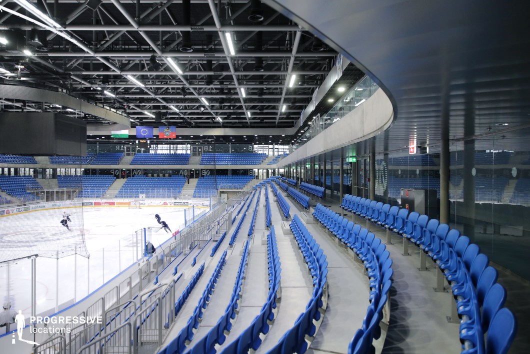 This image depicts a modern ice hockey arena featuring a sleek, enclosed design with strong architectural lines and high functionality. The central element is the ice rink, enclosed by transparent barriers, and surrounded by steeply tiered rows of blue plastic seats that curve gently with the shape of the arena. The composition emphasizes order and symmetry, with clear sightlines and unobstructed visibility from all seating levels. The ceiling structure is open and industrial, showcasing exposed grid beams, suspended lighting systems, and various mechanical elements in a clean, organized layout. Materials such as polished concrete, steel, and glass dominate the space, reinforcing the arena’s contemporary, utilitarian character. A glass-walled corridor above the seating hints at additional premium or media zones. The angle of the image, taken diagonally from the side stands, offers a dynamic sense of perspective, drawing the viewer’s eye toward the ice surface and scoreboard at the far end. The color palette is predominantly cool, with varying shades of blue and grey contributing to a crisp, technical aesthetic. This venue is particularly well-suited for productions requiring an enclosed sports environment with a clean, modern look, and is ideal for dynamic tracking shots, crowd simulations, or scenes emphasizing precision and energy.