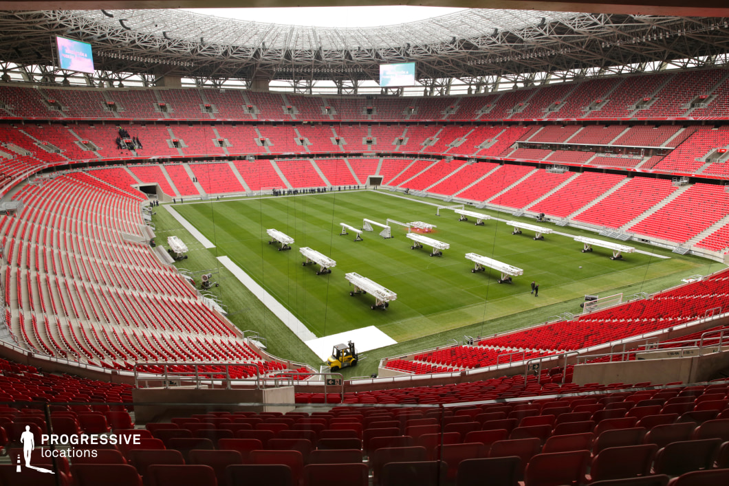This image depicts a large-capacity, modern stadium with a symmetrical, oval-shaped layout, ideal for large-scale events and dynamic aerial shots. The central field is composed of well-maintained green grass, currently set up with modular structures that suggest preparation for a non-sporting event, such as a concert or public gathering. The surrounding grandstands are filled with uniformly arranged red plastic seats, generating a visually striking contrast with the green of the field and reinforcing a strong geometric cohesion throughout the venue. The stadium features three continuous tiers of seating, reflecting an architectural emphasis on vertical layering and mass capacity. Steel and concrete dominate the structural materials, with a high, intricate metal roof canopy partially covering the uppermost seating levels. The open-air design above the field maintains natural lighting while the roof’s lattice structure adds both aesthetic and functional complexity. The photo is taken from a slightly elevated, central rear viewpoint, offering a panoramic view of the venue’s scale and form. The perspective highlights the radial symmetry and spatial rhythm of the seating rows, as well as the stadium’s potential for hosting cinematic scenes with grand visual scale. This location is well-suited for projects requiring a dramatic, high-energy atmosphere or settings with monumental scale and clean, modern design.
