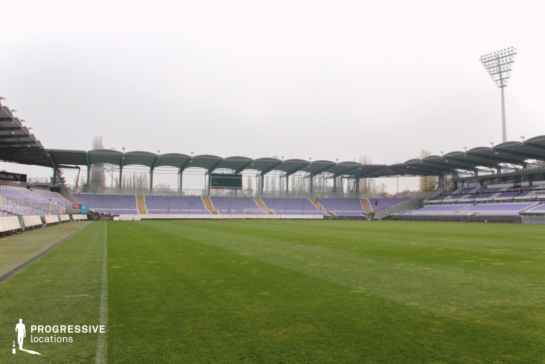 This image presents a medium-capacity football stadium with gently curved metal roofing and symmetrical, segmented stands clad in lavender and white seating. The architecture features an open design with visible structural framing and evenly distributed roof segments supported by slender steel columns. The field is natural grass, well-maintained, and enclosed by low walls and advertising panels. The setting evokes a functional, community-oriented sports venue suitable for both regional games and professional events. The consistent color palette, compact scale, and modest structural lines support versatile filming or photography use, particularly where a realistic, grounded stadium atmosphere is required.