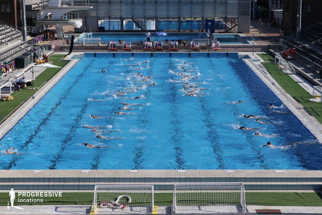 A large-scale outdoor Olympic swimming pool set in a professional training environment, equipped with multiple lanes and surrounded by tiered seating and sports infrastructure. The pool is actively used by a synchronized group of swimmers, emphasizing its function as a high-capacity training or competition venue. The facility features artificial turf around the perimeter, poolside sports gear storage, and a backdrop of additional training pools and diving platforms. The symmetrical layout, clean lines, and competitive ambiance make this location ideal for sport-themed productions, commercial shoots, or dynamic action sequences. Its versatility and well-maintained surroundings support both wide establishing shots and close-up, energetic scenes.