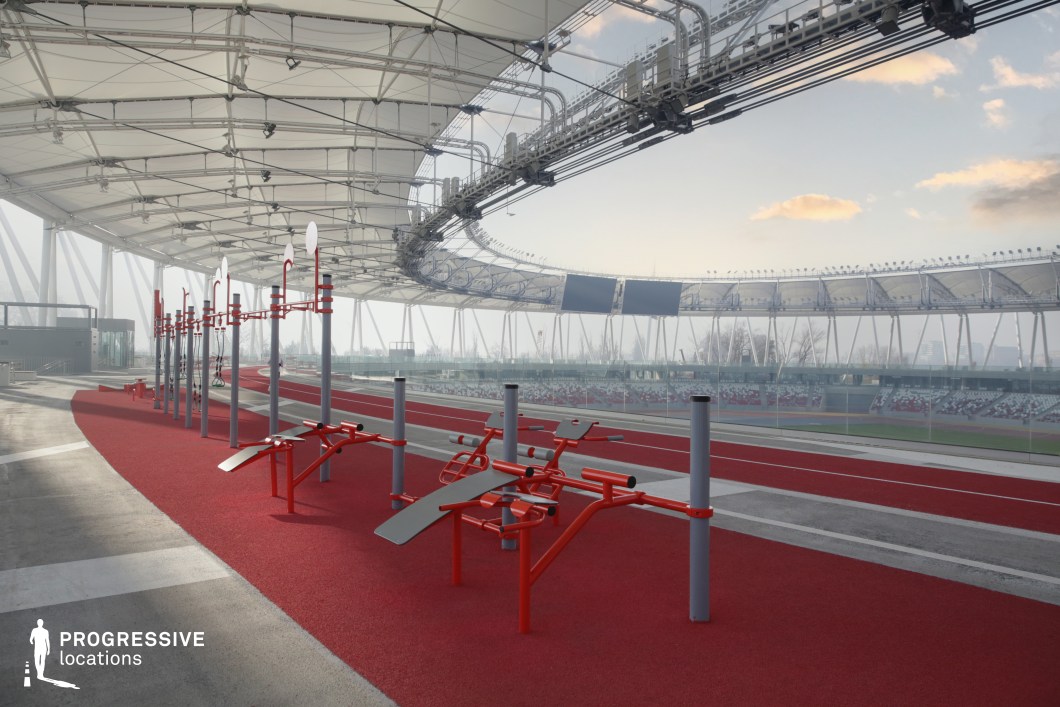 This visually striking image depicts a modern outdoor athletics facility featuring a dedicated training area under a large tensile canopy. The foreground is dominated by a series of fixed fitness and calisthenics equipment, constructed in red and silver metal and arranged on a bright red rubberized surface. These elements create a high-contrast, structured environment geared toward warm-up, strength, or conditioning exercises adjacent to a running track. The backdrop reveals the broader stadium layout, with spectator stands in red and white seating and a transparent perimeter barrier offering uninterrupted views. Above, the tensile roof structure is supported by slender white columns and crisscrossing cables, integrating lighting and electrical systems. The design emphasizes lightness and spatial openness while providing partial shelter and a visually dynamic overhead framework. Shot from a low diagonal angle, the image highlights both the functionality and modern aesthetics of the facility. The repetition of vertical poles, sweeping lines of the track, and layered sightlines toward the seating area contribute to a strong compositional rhythm. This location is ideal for sports-themed productions, fitness branding, or any creative concept requiring a contemporary, open-air setting with architectural clarity and vibrant spatial design.