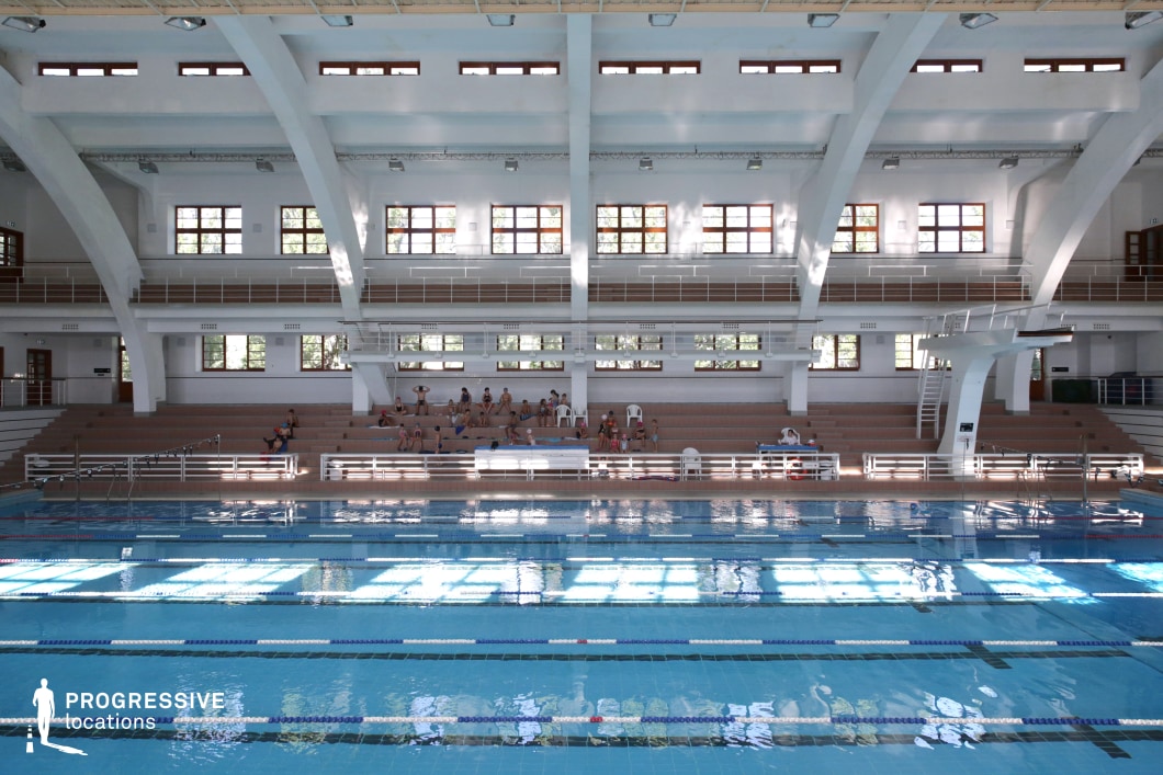 This traditional indoor swimming hall features a rectangular pool with clearly marked lanes and calm, reflective water, enclosed by a white structural framework with thick curved beams. The design incorporates a two-level layout with symmetrical balconies, wide stair access, and tiered beige seating that offers a subtle contrast against the predominantly white interior. Natural light floods the space through large wooden-framed windows positioned on both levels, creating bright, grid-like reflections on the pool surface. A diving platform is integrated on the right side, adding versatility to the facility. The overall setting blends classic architectural lines with soft modern updates, making it suitable for period or contemporary scenes requiring a serene and geometrically balanced aquatic interior.