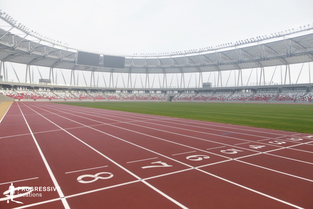 This image highlights a state-of-the-art athletics stadium from the perspective of the starting lanes on a red synthetic track. The viewpoint, low and angled, emphasizes the linear precision of the marked lanes and the wide, expansive design of the venue. The vibrant red track surface contrasts sharply with the green central field, creating a visually balanced and professional-grade sporting environment. The grandstands, featuring a red-and-white seating pattern, surround the track in a continuous elliptical arrangement. The structural canopy is supported by slender vertical columns and integrated lighting systems, enhancing both the architectural clarity and the technical sophistication of the venue. The roofing design remains elevated and visually light, allowing ample exposure to the sky while maintaining a sense of enclosure. The visual composition underscores the stadium's geometric coherence, drawing attention to its symmetry, depth, and functional zoning. The arrangement is ideal for large-format filming or photography, particularly for sequences that require uninterrupted perspective lines or wide horizontal space. This stadium offers a dynamic and flexible backdrop, especially suited to projects focused on themes of performance, competition, or modern infrastructure.