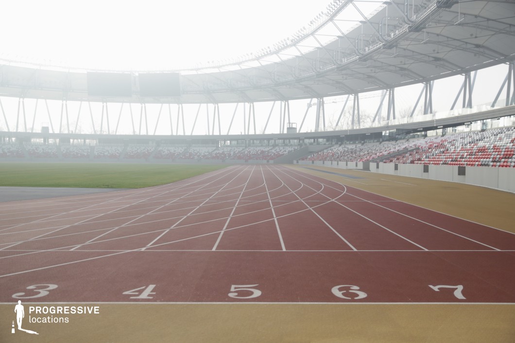 This image captures a sweeping view of a professional athletics stadium, focused on the numbered starting lines of a red synthetic running track. The gently curving lanes guide the eye into the spacious center of the venue, framed by structural columns and a tensile canopy above. The red and white seating pattern in the grandstands adds rhythmic visual texture, while the open design allows for soft ambient light to filter through the space. The camera angle is ground-level and slightly off-center, emphasizing both depth and motion, ideal for compositions involving dynamic forward movement. The surface of the track appears meticulously maintained, with lane markings crisply delineated in white against the red base. Architecturally, the stadium combines exposed steel frameworks with lightweight roofing, creating a balance between openness and enclosure. The visual symmetry and expansive lines make this setting ideal for film sequences, commercial shoots, or visual narratives requiring a contemporary, action-ready sports environment.