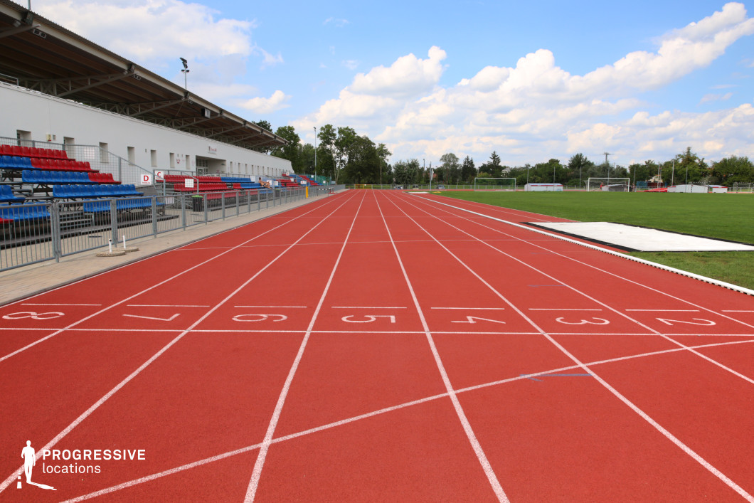 This image captures the straight segment of a red synthetic running track, seen from a low, central perspective, emphasizing the neatly marked white lane numbers from 1 to 8. The track runs parallel to a small grandstand area with simple, metal-framed bleachers featuring blue and red seating. Behind the stands, a white, linear facility building with a sloped roof adds a minimal architectural backdrop. On the right side, a well-maintained green grass field borders the track. The setting provides a clean, symmetrical visual ideal for commercial or sports-themed productions needing a straightforward athletics venue with a modest, accessible character.