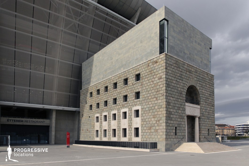 This location features a compact, fortress-like stone pavilion set against the backdrop of a contemporary stadium. The structure showcases a combination of heavy stone masonry on the lower levels and smoother, modern cladding on the upper portion. The main façade is characterized by a series of small, evenly spaced square windows and a dominant arched entrance with neoclassical columns beneath, suggesting historical or ceremonial significance. The blend of modern stadium infrastructure and robust classical forms creates a unique visual contrast. The setting is completed by a broad, paved plaza that allows for wide camera angles and large-scale event setups. The building’s textures, symmetry, and interplay of light on the stone surfaces make it an ideal location for stylized productions requiring architectural gravitas and urban formality.