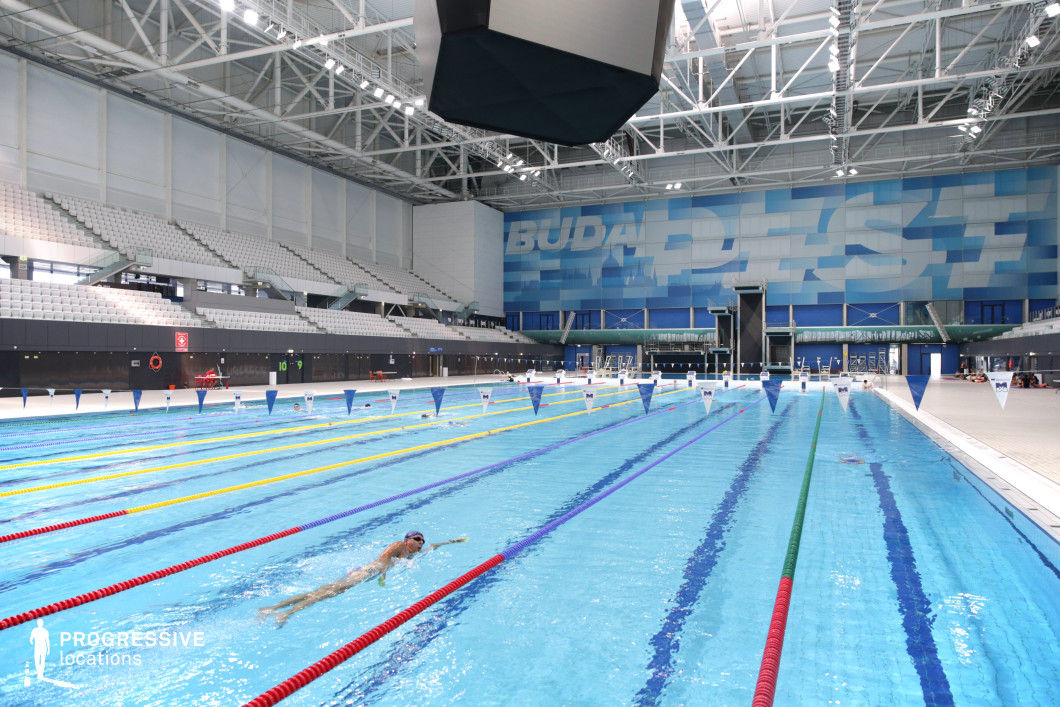 This high-capacity indoor swimming arena features a full-length competition pool designed for professional aquatic events. The image captures the pool from a slightly angled perspective, emphasizing the spatial depth and the structure’s symmetry. Multiple lanes, separated by colored dividers, stretch toward a large blue-toned wall adorned with stylized typography, reinforcing the venue’s identity and adding a visually dynamic backdrop. The architectural design combines white and blue hues with industrial elements. Overhead, a robust steel truss system supports integrated lighting and an oversized, suspended video scoreboard. The spectator stands are clad in uniform white seats arranged in ascending tiers, offering clear views from every angle. Smooth tiled surfaces and metal railings enhance both the aesthetic and functional aspects of the space. This venue’s clean, contemporary appearance and large-scale proportions make it particularly suitable for productions requiring a sleek, high-performance environment. Its structural openness and excellent visibility support a wide range of creative uses in sports, commercial, or high-tech contexts.