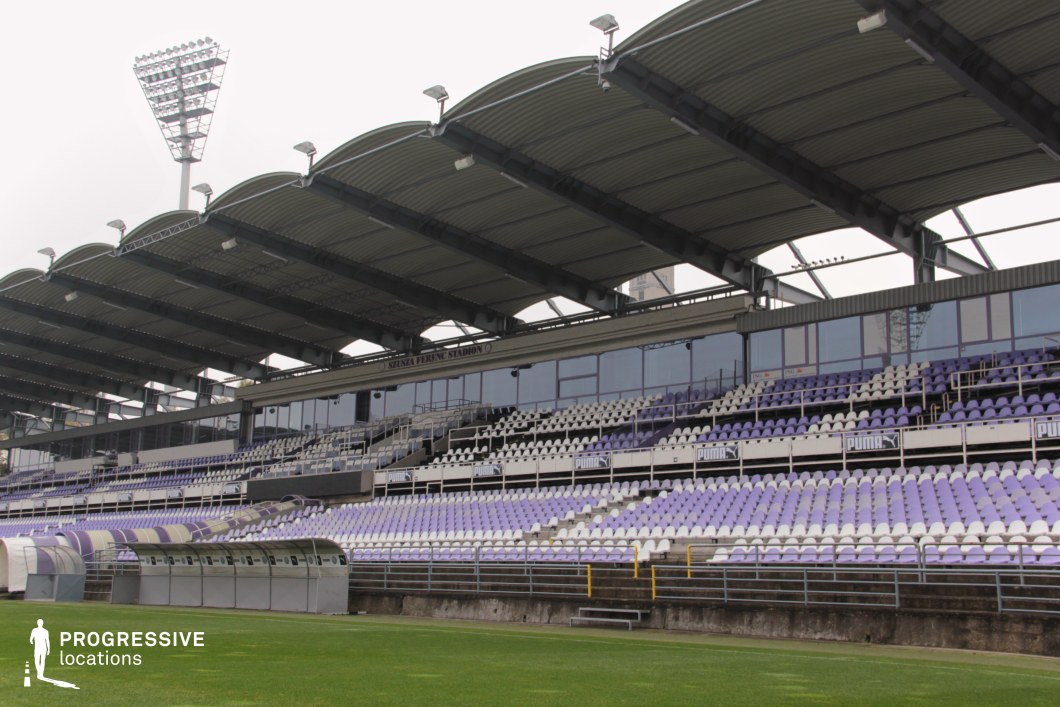 This stadium grandstand showcases a compact yet structured layout, characterized by alternating rows of white and pale purple plastic seating beneath a repeating arched metal canopy. The stand includes a glass-fronted central section, likely reserved for VIPs or media, flanked by standard spectator areas. Advertising panels and branded barriers add a commercial sporting context. Constructed from concrete and steel, the stand conveys a solid, pragmatic design. The modest scale, clean sightlines, and visually consistent seating pattern make it ideal for productions focusing on local matches, training sessions, or community-level sports events in an urban setting.