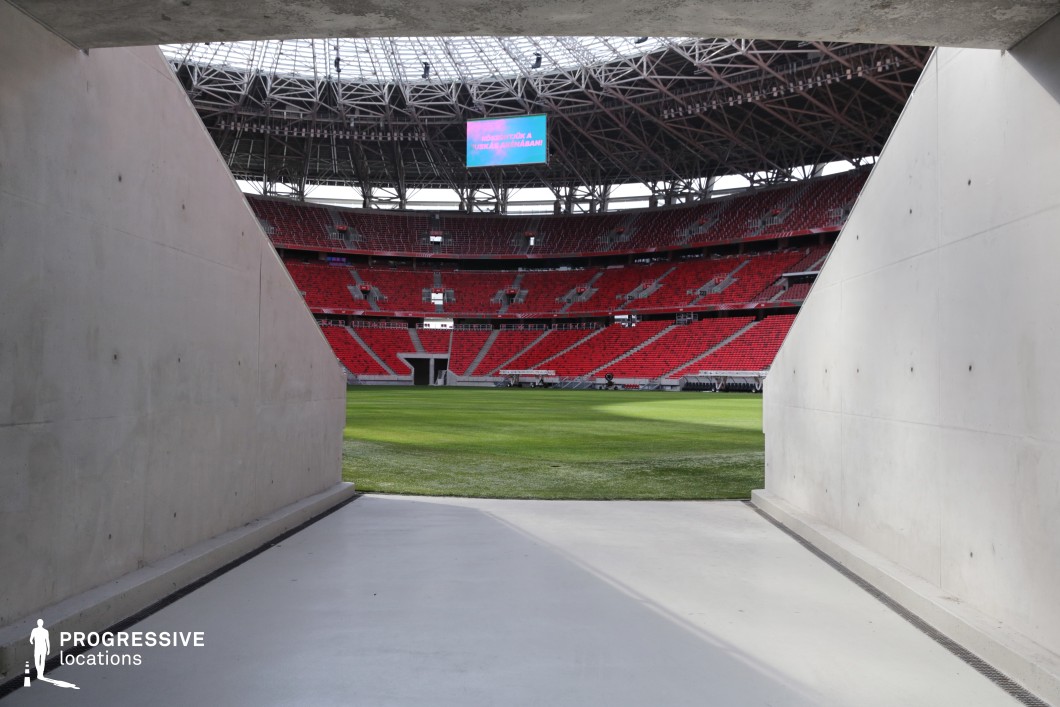 This image offers a dramatic perspective from within a concrete player tunnel, opening directly onto the vibrant green field of a large-scale football stadium. The symmetrical framing of the smooth concrete walls creates a stark visual contrast with the burst of color and architectural complexity revealed beyond. Red tiered seating, steel roof trusses, and a centrally suspended LED screen dominate the background, reinforcing the stadium’s scale and modern infrastructure. The slightly lowered vantage point emphasizes the transitional nature of the tunnel—evoking movement from behind-the-scenes preparation into the spotlight of ompetition. The field’s edge is marked by a slight shadow cast by the stadium’s roof, which adds dimensional depth and natural mood to the otherwise clean architectural lines. This location is perfectly suited for narrative transitions, athlete walk-outs, or symbolic moments of emergence and anticipation in film or commercial contexts. Its powerful geometry and clear function make it an instantly recognizable and versatile visual motif.