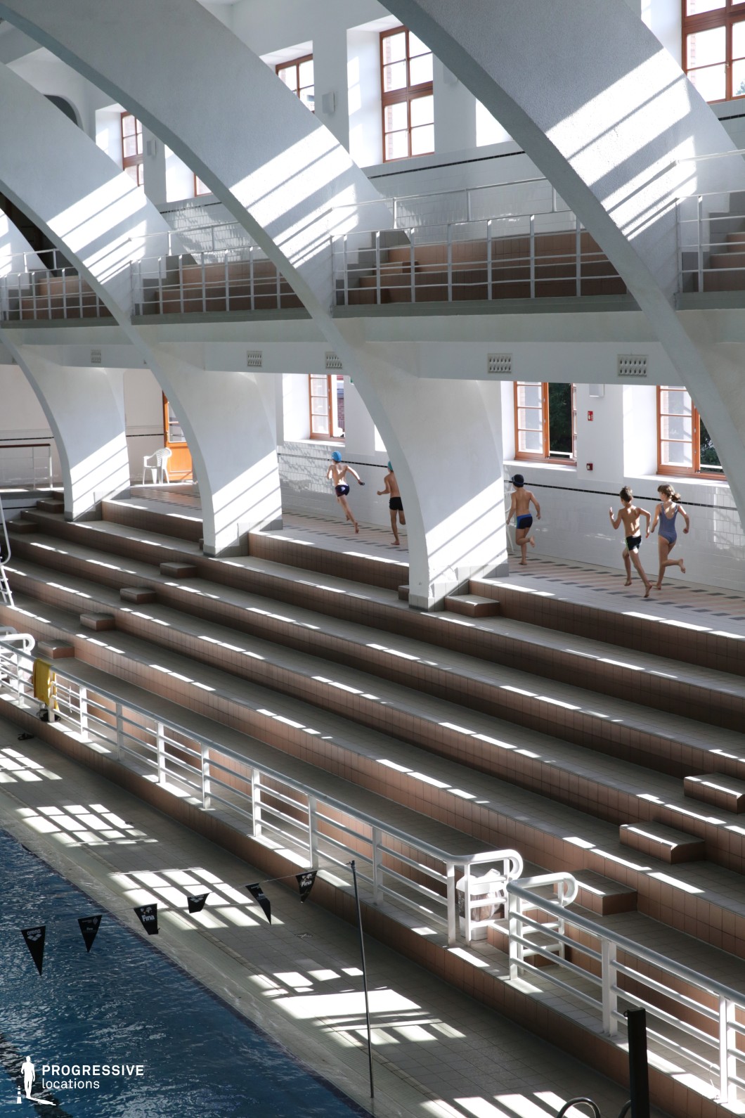 This indoor pool facility features a striking arched beam structure that creates a rhythmic architectural pattern above a terraced seating area. The wide, tile-covered steps serve both as spectator benches and transition levels leading down to the pool. The space is defined by a warm, natural palette—light brown tiles, white structural elements, and wooden-framed windows that invite ample daylight. The vertical and horizontal lines formed by railings, steps, and shadows contribute to a visually structured yet dynamic environment. The setting is ideal for scenes focusing on sports training, movement, or rhythm, particularly in aquatic or athletic contexts that benefit from classical design and clean, modern lighting.