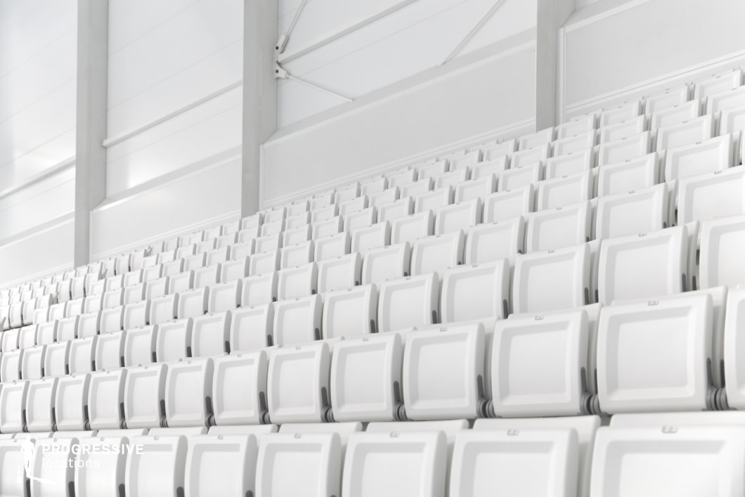 This close-up image focuses on a section of modern, tiered stadium seating arranged in a clean, repetitive pattern. The chairs are uniform in design, featuring white plastic backs with soft grey structural elements, contributing to a highly minimalistic and sterile visual character. The setting suggests a newly constructed or recently renovated indoor sports facility. The image is taken from a slightly angled side perspective, emphasizing the rhythmic geometry and ascending alignment of the seats. The background structure is composed of light grey and white architectural surfaces with visible panel joints and vertical supports, reinforcing the orderly, modular nature of the venue. This location is particularly suited for productions or campaigns requiring a crisp, neutral environment with architectural symmetry and a clean design language. The visual uniformity and neutral color palette make it an adaptable backdrop for various commercial or cinematic applications.