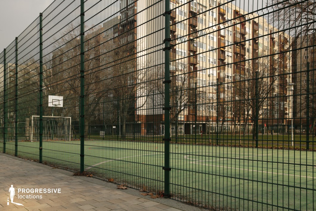 A fenced multi-use sports court situated in a densely built urban residential area, surrounded by high-rise apartment blocks. The court surface is made of green synthetic turf, and it features both a basketball hoop and a football goalpost. The tall, dark green metal mesh fence encloses the entire area, providing a sense of containment and privacy, while allowing visibility through to the surrounding environment. The backdrop of concrete residential towers and bare trees emphasizes a gritty, authentic urban mood. This location is ideal for contemporary youth culture visuals, community-centered storytelling, or productions needing a versatile inner-city sports setting.