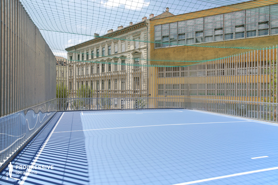 This elevated sports court features a bright blue modular surface enclosed by vertical metal slats and a protective net overhead. The minimalist design and sharp white line markings lend the space a crisp, controlled aesthetic. The fencing allows for filtered views of the surrounding architecture, contributing depth and rhythm to the composition through light and shadow play. In the background, contrasting architectural styles define the location’s character. A neoclassical building with ornate stone façades and rhythmic window arrangements stands adjacent to a modernist yellow structure with grid-like window patterns, creating a dynamic visual dialogue. The juxtaposition highlights the blend of historical and contemporary urban fabric. This rooftop court offers a highly stylized and versatile filming environment, ideal for productions seeking a blend of modern design elements with urban heritage in a spatially compact, elevated setting.