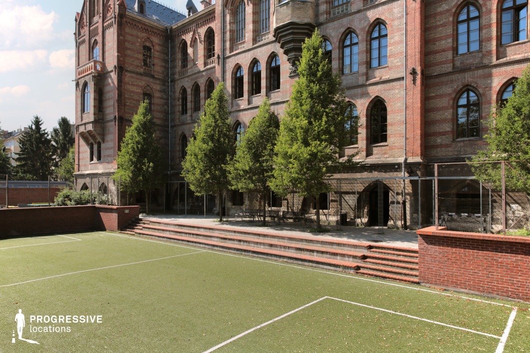 This enclosed green courtyard is part of a striking historic brick building, likely a former school or institutional structure with Neo-Gothic architectural details such as pointed arch windows, ornamental cornices, and a turreted corner. The field itself features an artificial grass surface with white line markings, suitable for recreational sports or school activities. Surrounding the courtyard is a red brick retaining wall with a wide stairway leading to a shaded patio lined with mature trees and outdoor seating. The visually rich setting offers a unique contrast between the formality of the historic architecture and the functionality of a modern play or sports area, making it a distinctive backdrop for period-inspired urban scenes or educational-themed productions.