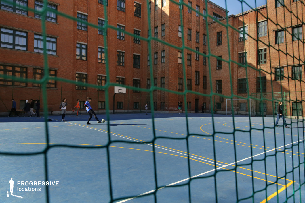 This compact urban courtyard is enclosed by a uniform red-brick school building featuring dense window grids and decorative brick detailing. At its center lies a blue multi-use sports court, clearly marked with white and yellow lines for basketball and soccer, and partially viewed through a green protective netting that adds a textured foreground layer. The built environment is consistent in style, with repetitive rectangular window patterns and solid vertical surfaces that emphasize symmetry and architectural cohesion. The high brick walls surrounding the court create a secluded, acoustically contained space, well-suited for private or institutional activities. The perspective through the net enhances the sense of enclosure and realism, making this site particularly appealing for educational, youth-oriented, or retro urban visuals requiring an authentic, grounded school setting.