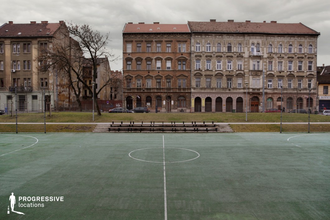 This location centers on a rectangular outdoor sports court with faded green synthetic surfacing and clearly marked white boundary lines, flanked by a simple wooden grandstand. The court is set in a flat urban park space, bordered by a narrow grassy area and paved walkways that emphasize its accessibility and public use function. The background is visually defined by a row of historic, late 19th to early 20th century residential buildings, characterized by ornate plasterwork, arched ground-level arcades, and varied window rhythms across four stories. The façades exhibit a weathered, textured patina that enhances the location’s lived-in, East-Central European atmosphere. Subtle architectural contrasts between the buildings create a rhythm of variation while maintaining a cohesive historic aesthetic. The symmetrical composition and open perspective make this site ideal for static wide shots or dynamic sports-related visuals, while the architectural backdrop provides a strong sense of urban realism and period character.
