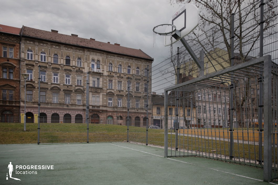 This modestly sized urban sports court is surfaced in muted green and enclosed by a full-height metal fence. A weathered basketball hoop and metal-framed soccer goal occupy the foreground, indicating the court's dual-function design. The court sits slightly below street level, bordered by a sloped grassy embankment, contributing to a compact, enclosed atmosphere. The backdrop is formed by a row of richly detailed late 19th to early 20th-century residential buildings. Their façades display a mix of light brickwork, arched windows, ornate cornices, and ground-level arcades, adding strong architectural texture and depth to the setting. The combination of historical urban architecture with a utilitarian recreational space offers an authentic, slightly gritty setting suited for neighborhood-centric narratives, youth culture visuals, or productions exploring the intersection of public space and residential environments.