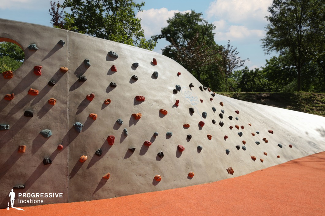 The image captures a modern outdoor bouldering wall integrated into a landscaped park setting. The concrete climbing surface is organically shaped, with a smooth curvature and embedded multi-colored climbing holds arranged in varying difficulty. The orange rubberized flooring ensures safety and adds a vibrant visual contrast to the neutral wall surface. Surrounding trees and scattered natural shadows enhance the environment with a recreational and family-friendly ambiance. This compact, sculptural feature is ideal for urban sports shoots, active lifestyle content, or promotional visuals emphasizing inclusive outdoor play and contemporary park design.
