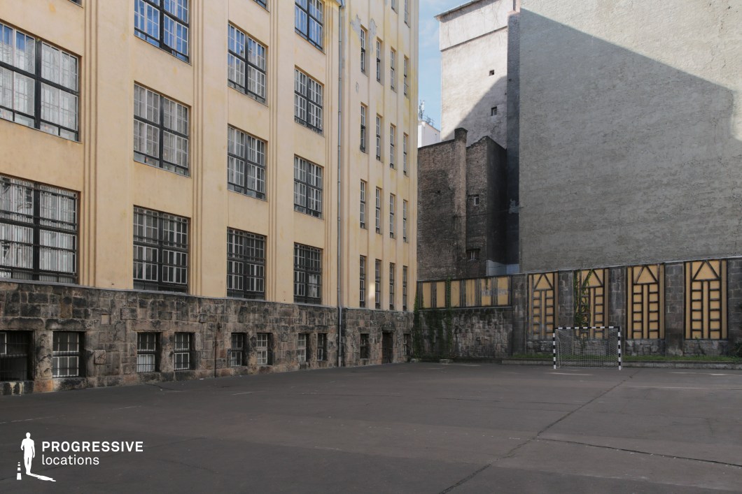 This enclosed urban courtyard features a bare asphalt surface with a small handball goal positioned against a weathered stone and stucco wall. The space is framed on one side by a tall institutional building, likely an old school or public facility, with a façade of soft yellow tones and vertical bands of industrial-style windows protected by grilles. The opposite boundary reveals a layered architectural background of aged brick and concrete walls, showcasing a range of surface textures—from patched masonry to stark grey façades. The walls are punctuated with tall, narrow decorative window insets, bordered by climbing ivy, adding a historic and slightly dramatic visual element to the setting. This space exudes an atmospheric, almost theatrical quality, ideal for gritty urban storytelling, reflective scenes, or low-key sports activities in a closed, semi-industrial environment. Its rawness and spatial intimacy suit productions looking for authentic, historic, or institution-adjacent locations.