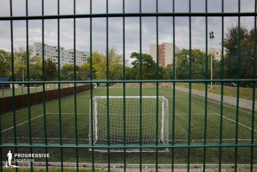 This rectangular artificial turf soccer field is framed by tall green fencing and low concrete retaining walls, creating a self-contained recreational zone. The goalpost is centrally positioned in the foreground, viewed through vertical metal bars that add a strong sense of depth and structure to the composition. Beyond the pitch, the background is defined by mid-century residential buildings, typical of Central and Eastern European housing estates. Their repetitive façades and muted tones contrast with the surrounding vegetation, including mature trees and manicured grass. Tall floodlights suggest nighttime usability and underline the space’s public-sports functionality. This location offers a realistic, grounded atmosphere suited for productions requiring an everyday urban backdrop with clear visual separation between athletic space and dense residential environments.