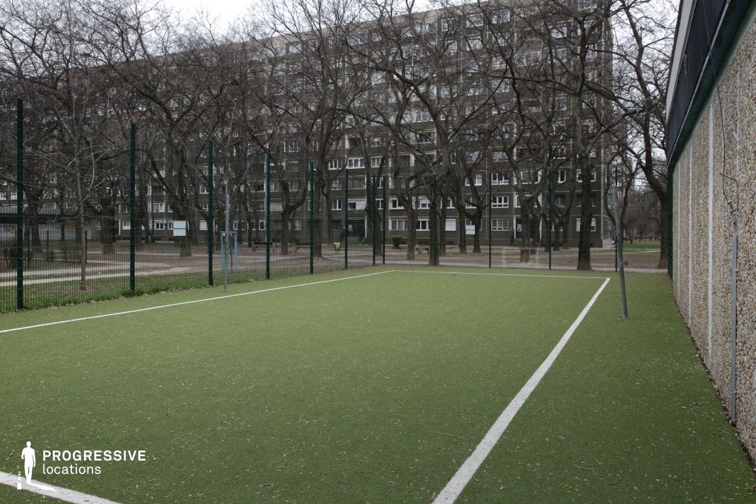 This compact outdoor sports field is defined by artificial green turf and bordered by white painted lines, creating a clean and structured space ideal for recreational use. A tall metal fence encloses the court on all visible sides, reinforcing a sense of separation from the surrounding environment. One wall is covered with pebble-dashed concrete panels, contributing to the utilitarian atmosphere. In the background, rows of leafless trees partially obscure a large residential block, characterized by its repetitive window patterns and muted tones. The field is nestled within a broader urban park setting, visible through the fencing, and connects with walkways and benches beyond its boundaries. This location offers a stark, minimalist sports environment, perfect for urban scenes that require a contrast between recreational and residential elements, or for storytelling rooted in city life and community spaces.