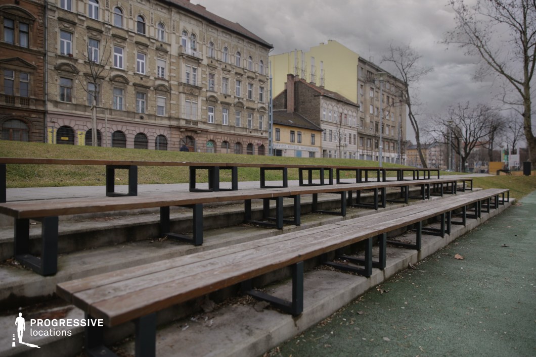 This outdoor location features a set of stepped wooden bleachers with metal supports, integrated into a sloped concrete base. The seating arrangement faces a green park area and is positioned along a paved recreational surface, suggesting its use for observing adjacent sports or community events. In the background, a row of ornate, early 20th-century residential buildings establishes a strong urban character. The facades exhibit historicist architectural details such as arched windows, stone cornices, and decorative brickwork, contributing to a textured and layered streetscape. The subtle variation in color and style across the buildings adds visual rhythm while maintaining overall cohesion. This site offers a compact and atmospheric setting ideal for urban community-focused scenes. The combination of classic architecture and practical public infrastructure creates a visually rich environment adaptable to sports, lifestyle, or documentary-style productions.