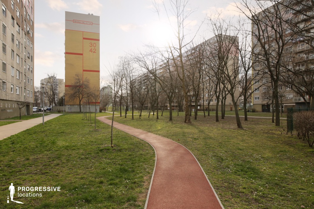 An urban green zone featuring a gently curving red jogging or walking path set between tall, weathered apartment buildings. The space includes grassy lawns, young and mature trees, and minimalistic streetlights, forming a balanced interface between built environment and nature. The architecture is dominated by mid-to-late 20th-century concrete residential towers, with one facade marked by a bold numerical mural for visual emphasis. This location provides a subtly nostalgic atmosphere, ideal for scenes requiring an everyday, lived-in urban park setting. Its linear perspective and mix of soft vegetation and raw architecture make it adaptable for narrative, fashion, or lifestyle productions.