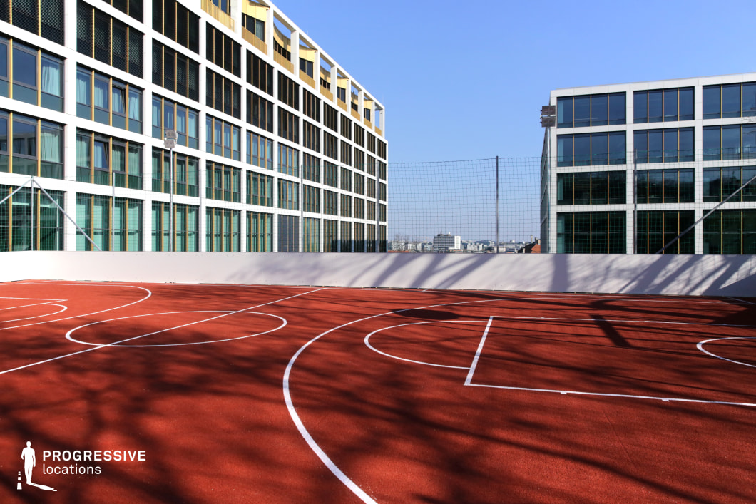 This elevated sports location features a vibrant red rooftop court designed for basketball, enclosed by a solid white perimeter wall and protective netting. The surface is clean and uniformly marked with white lines, creating a strong contrast with the surrounding architecture. Flanking the court are contemporary office or institutional buildings with modular facades defined by white grid frameworks and large rectangular windows. Their repetitive geometry, cool-toned glass, and metallic accents reinforce the site’s modern, minimalist aesthetic. The view extends toward a distant urban skyline, contributing a sense of openness despite the enclosed structure. This location offers a clean, high-contrast visual setting suitable for stylized, futuristic, or corporate-urban productions. Its elevated perspective and architectural precision make it ideal for dynamic scenes with controlled backgrounds and a strong graphic quality.