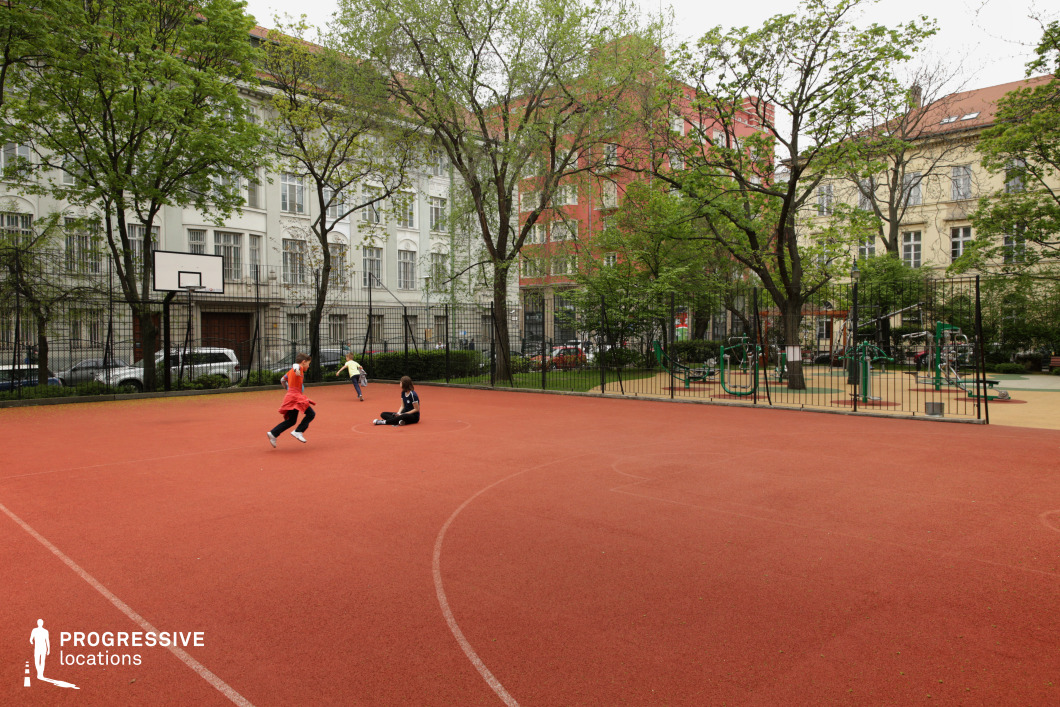 This urban sports court is situated within a residential neighborhood bordered by classic European apartment buildings and mature trees. The court is surfaced with a red rubberized material and marked for various ball games, making it suitable for school or community recreation. A single basketball hoop is mounted on a pole at one end, while the surrounding area is enclosed with black metal fencing for safety and containment. Adjacent to the court is an open-air fitness area featuring green and gray gym equipment. The overall atmosphere is vibrant and community-focused, ideal for urban activity scenes, schoolyard storytelling, or public space documentation.