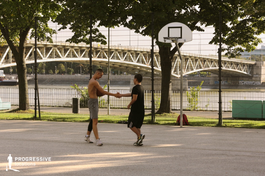This open-air basketball court is located alongside a riverside promenade, enclosed by metal fencing and shaded by mature trees. The playing surface is a light-toned, flat pavement rather than synthetic material, lending the court a minimalist, urban park feel. A simple, white backboard and hoop structure stands near the center of the frame. Behind the fence, a prominent steel truss bridge spans the river, adding architectural depth and visual dynamism to the composition. The bridge’s repetitive triangular geometry and the rhythmic vertical bars of the fence create layered patterns that enhance the image’s spatial depth. The river itself and the distant embankment serve as a tranquil visual base beneath the industrial structure. This location merges urban sport and riverside calm, offering an ideal setting for productions seeking contrast between built infrastructure and natural waterfronts within a metropolitan context.