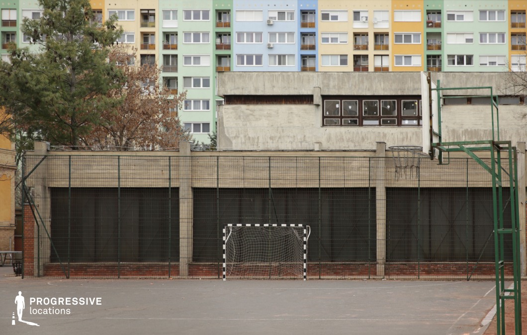 A modest asphalt sports court set against a backdrop of mid-century brutalist architecture and vibrant, multi-colored residential buildings. The court is equipped with a metal basketball hoop and a small handball goal, enclosed by high fencing. The background features faded concrete surfaces and a striking row of pastel-toned high-rise apartments, offering a dynamic contrast of raw texture and bright geometry. The location has a distinctly urban, retro aesthetic with authentic Eastern European tones. It's suitable for gritty youth culture scenes, nostalgic atmospheres, or productions that require a visually engaging contrast between utilitarian and playful elements.