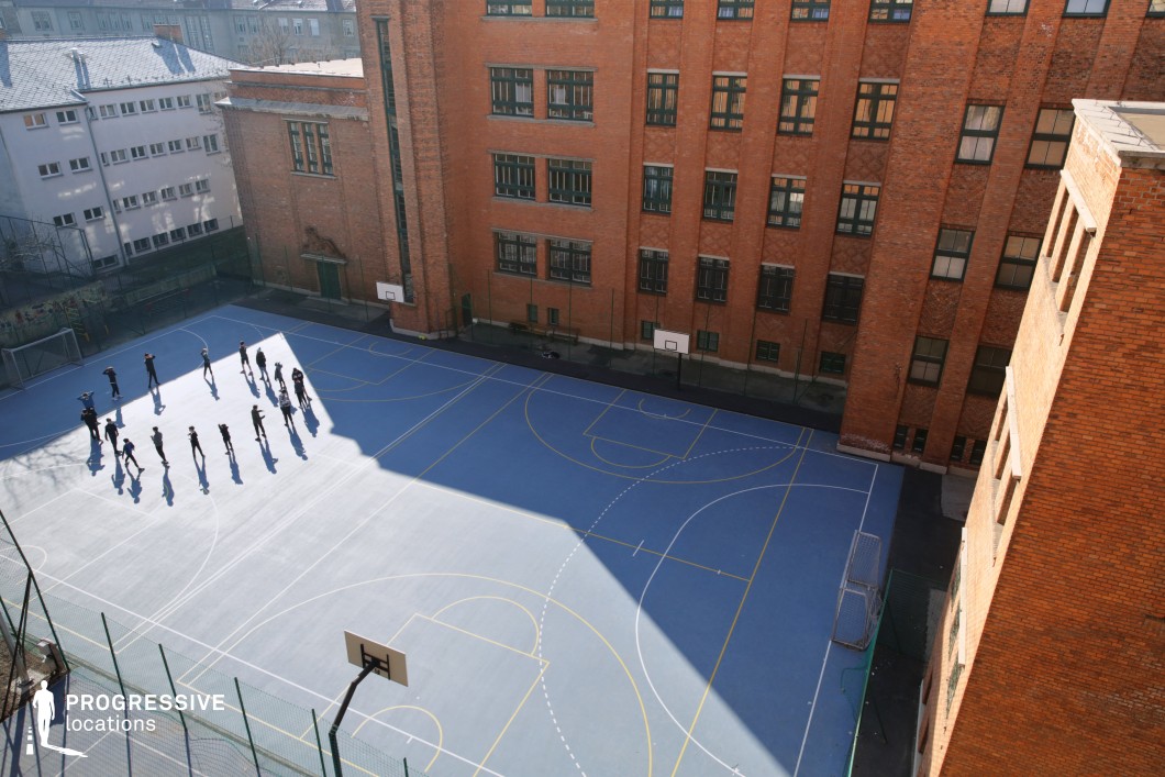 This enclosed courtyard sports field is bordered on three sides by tall institutional brick buildings, forming a rectangular, inward-facing environment ideal for school or recreational use. The playing surface is a matte blue court, clearly marked for multiple sports, including basketball and soccer, with crisp white and yellow lines. The surrounding architecture features industrial-style red brickwork, large multipane windows, and minimal decorative detailing, evoking early-to-mid 20th-century school or public building design. The space is visually segmented by light and shadow due to the high surrounding walls, adding strong geometric contrast to the composition. Viewed from an elevated angle, the court’s symmetry and enclosed nature are emphasized, making this location particularly suitable for scenes requiring structured, institutional atmospheres or controlled, private activity zones in a dense urban context.