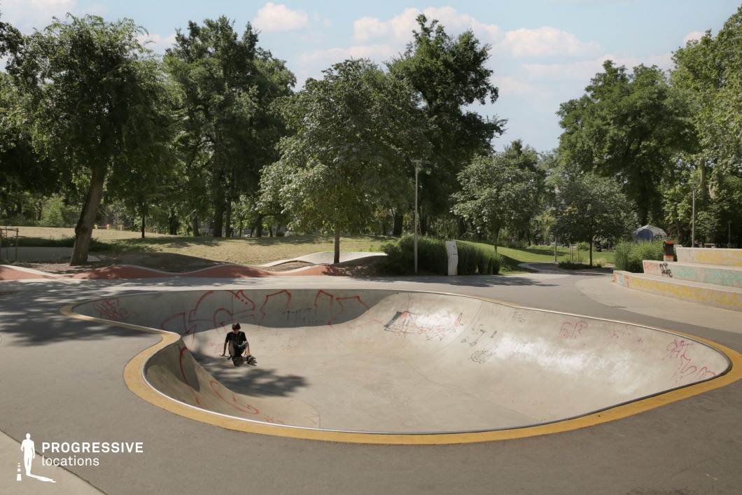 This location features a modern outdoor skatepark centered around a large, smoothly contoured concrete bowl. Its organically flowing shape is bordered with a yellow edge marking and adorned with faint graffiti in red and black tones. The skatepark is set within a spacious, tree-filled park, surrounded by lush greenery and meandering paths that provide depth and natural framing to the scene. The expansive, unobstructed surfaces and surrounding open space make this site particularly adaptable for dynamic action shots, youth culture themes, or lifestyle visuals. The balance between urban edge and natural backdrop enhances its suitability for versatile creative applications.
