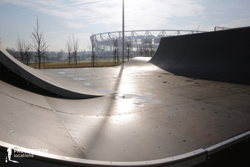 A sleek metal half-pipe and adjoining ramps form the core of this minimalist skate park, positioned in an expansive, open urban green space. The surface of the ramp is riveted and slightly reflective, with subtle water patches catching light and enhancing the textural detail. Sparse young trees line the background, while a large, modern stadium structure with a striking skeletal frame is visible in the distance, adding a sense of scale and futuristic contrast. The park’s flat layout, unobstructed views, and geometric curves make it ideal for dynamic urban-themed productions, youth culture content, or commercial shoots focusing on movement, energy, and street-style aesthetics.