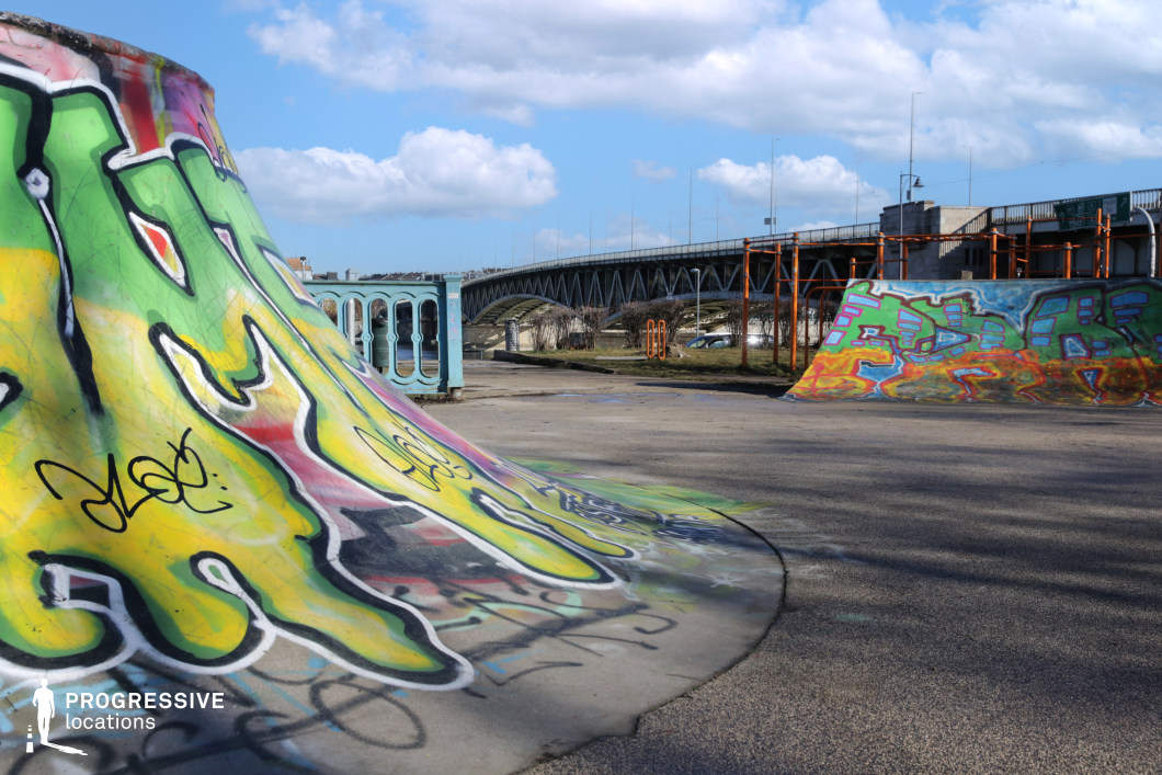 This open-air skatepark features vividly spray-painted ramps and halfpipes set against an industrial riverside backdrop. The concrete elements are fully covered in colorful graffiti art, lending the space an urban, raw, and expressive character. A long, arched bridge structure and retro-style railings mark the perimeter, while nearby orange metal installations add a layer of structural interest. The juxtaposition of bold street art, expansive paved surfaces, and a scenic riverside location makes this an ideal setting for dynamic youth culture, alternative fashion shoots, or music video scenes with a rebellious or energetic edge.