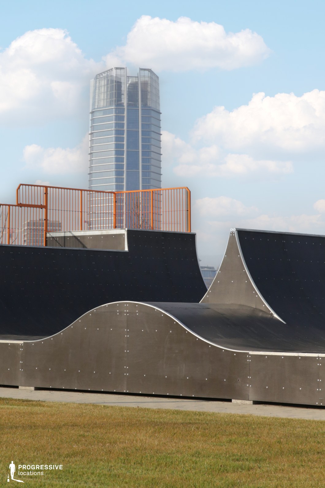 A sculptural outdoor skate ramp set against the backdrop of a striking, contemporary high-rise. The skatepark’s modular black surfaces and precision-riveted curves form a dynamic foreground, contrasted by the clean vertical lines of the reflective glass tower rising in the distance. Orange safety railings provide visual accentuation and frame the ramp structure with bold geometric lines. This location offers a visually compelling contrast between modern sports infrastructure and sleek urban architecture. Ideal for fashion editorials, lifestyle shoots, or action-driven scenes that call for a gritty yet highly graphic urban setting with a futuristic edge.