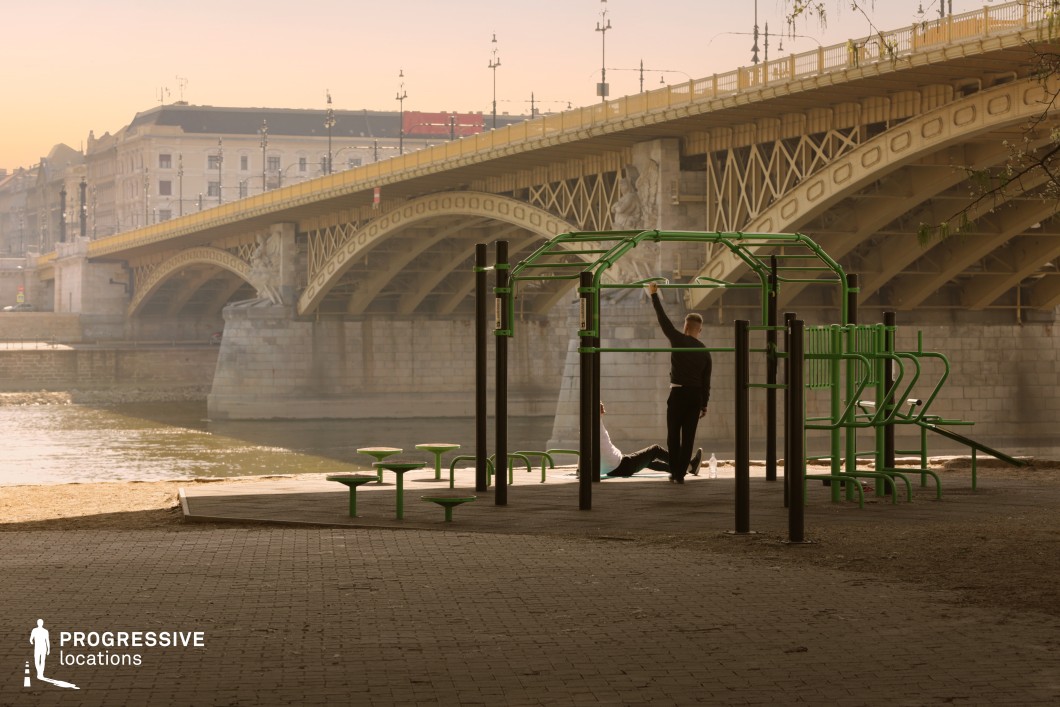 This location features a compact outdoor fitness park positioned directly along a riverside promenade, visually anchored by a large, historic iron bridge in the background. The foreground is dominated by a structured calisthenics workout area, incorporating sleek, green metal bars and stations installed on a dark rubberized base, framed by interlocking stone paving. The bridge beyond showcases ornate ironwork, massive stone pillars, and sculptural details, contributing a classic urban aesthetic. Architecturally, the bridge blends industrial and historic styles with its arched metal structure and detailed stone foundations. Surrounding buildings exhibit a mix of mid-rise, neoclassical facades, adding a refined, metropolitan atmosphere. The spatial composition contrasts the clean geometry of the exercise structures with the ornate curvature of the bridge, creating visual depth. Ideal for urban scenes that require a mix of contemporary function and historical urban context, this site provides a dynamic backdrop for fitness, lifestyle, or architectural-focused visuals.