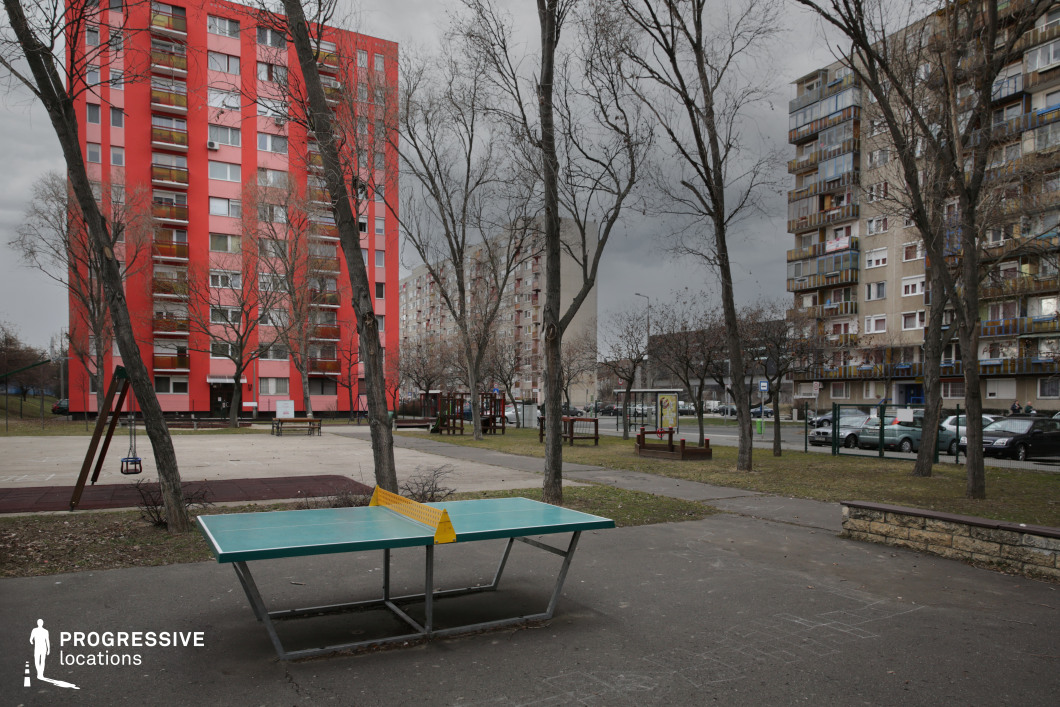 This suburban recreational space is anchored by a concrete table tennis table with a turquoise surface and yellow net, placed within a paved zone surrounded by sparse vegetation and bare trees. The setting includes a modest playground and walking paths, indicating its function as a shared public space. In the background, several residential high-rises create a dense architectural frame. The most visually prominent is a vivid red building with repetitive balconies, which contrasts sharply with the muted tones of the adjacent panel buildings. The mix of bright and neutral façades introduces a layered urban texture that reflects both renewal and the legacy of mid-century mass housing. The juxtaposition of communal sport infrastructure with post-socialist residential architecture makes this location ideal for productions exploring themes of neighborhood life, generational shifts, or youth activities in built-up suburban environments.