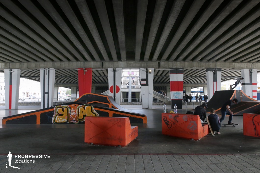 This dynamic location is a covered skatepark set beneath a large urban overpass. The space features various ramps, quarter pipes, and graffiti-tagged obstacles, with bold black and orange design elements adding a high-contrast visual identity. The concrete pillars supporting the overpass are painted in graphic patterns and national flag motifs, contributing to an edgy, metropolitan aesthetic. The raw concrete ceiling and exposed structural beams emphasize the industrial nature of the setting, while wide open sightlines allow visibility of distant staircases and passing foot traffic. The site blends architectural brutalism with youth-driven subculture, making it a vibrant yet gritty environment. Ideal for skate or street-culture themed productions, this location offers controlled lighting, bold lines, and authentic urban energy—perfect for music videos, lifestyle branding, or scenes centered on alternative sport and underground culture.