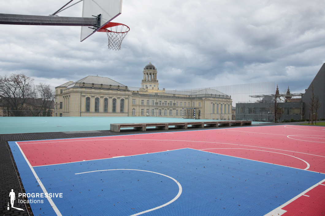 This striking location features a vividly colored rooftop sports court combining red and blue modular playing surfaces, outlined with white lines for basketball. The court is enclosed by sleek vertical metal fencing, providing a sense of safety without obstructing the view. Several minimalist concrete benches line the court, emphasizing its modern, urban design. In the background, a monumental neoclassical building dominates the visual field, characterized by large arched windows, light-colored stone walls, and a distinctive dome rising centrally. The architecture conveys institutional grandeur and historic sophistication, contrasting with the contemporary athletic setting. Additional structures with pointed spires and modern forms enhance the eclectic skyline and architectural layering. The elevated perspective, open composition, and visual tension between modernity and historical depth make this location highly adaptable for productions requiring an unusual mix of athletic, urban, and academic or cultural environments.
