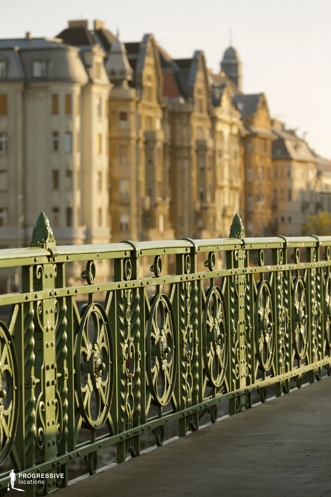 A decorative green iron bridge railing with intricate circular and geometric patterns dominates the foreground, exhibiting detailed craftsmanship and an old-world charm. The metal structure is solid yet elegant, adorned with rivets and ornamental finials, contributing to a sense of historical significance. In the softly blurred background, a row of grand, early 20th-century buildings with elaborate facades and domed rooftops creates a visually rich urban setting. The warm, muted tones of the architecture contrast with the cool green of the railing, adding depth to the composition. This location is ideal for filming period dramas, romantic narratives, or historical thrillers, offering a European-style ambiance suited for atmospheric cityscape shots or introspective character moments.