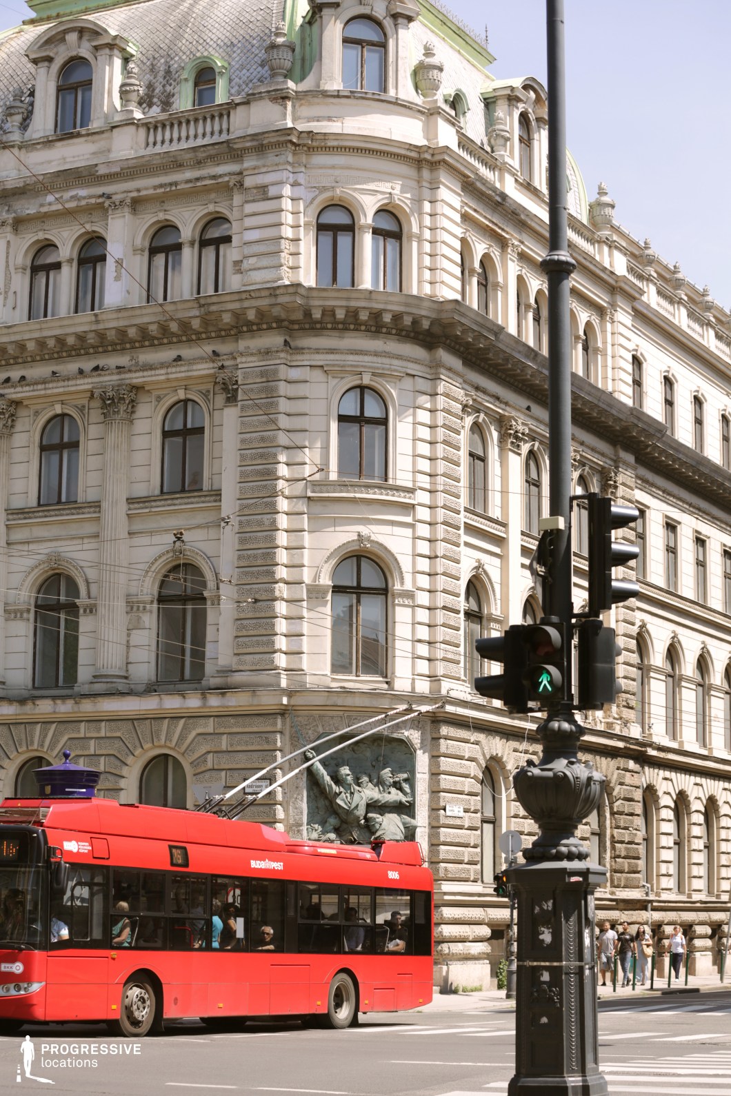 This image features a stately historic corner building with ornate neoclassical architecture, characterized by arched windows, decorative stonework, and a mansard-style roof with green copper accents. A sculptural relief on the facade adds a dramatic element, reinforcing the building’s historical significance. The street scene is marked by a red trolleybus and an ornate lamppost with a pedestrian crossing signal, emphasizing the urban environment. The location’s grandiosity and European city aesthetic make it ideal for filming period dramas, political thrillers, or scenes depicting metropolitan sophistication, historical intrigue, or bustling city life.