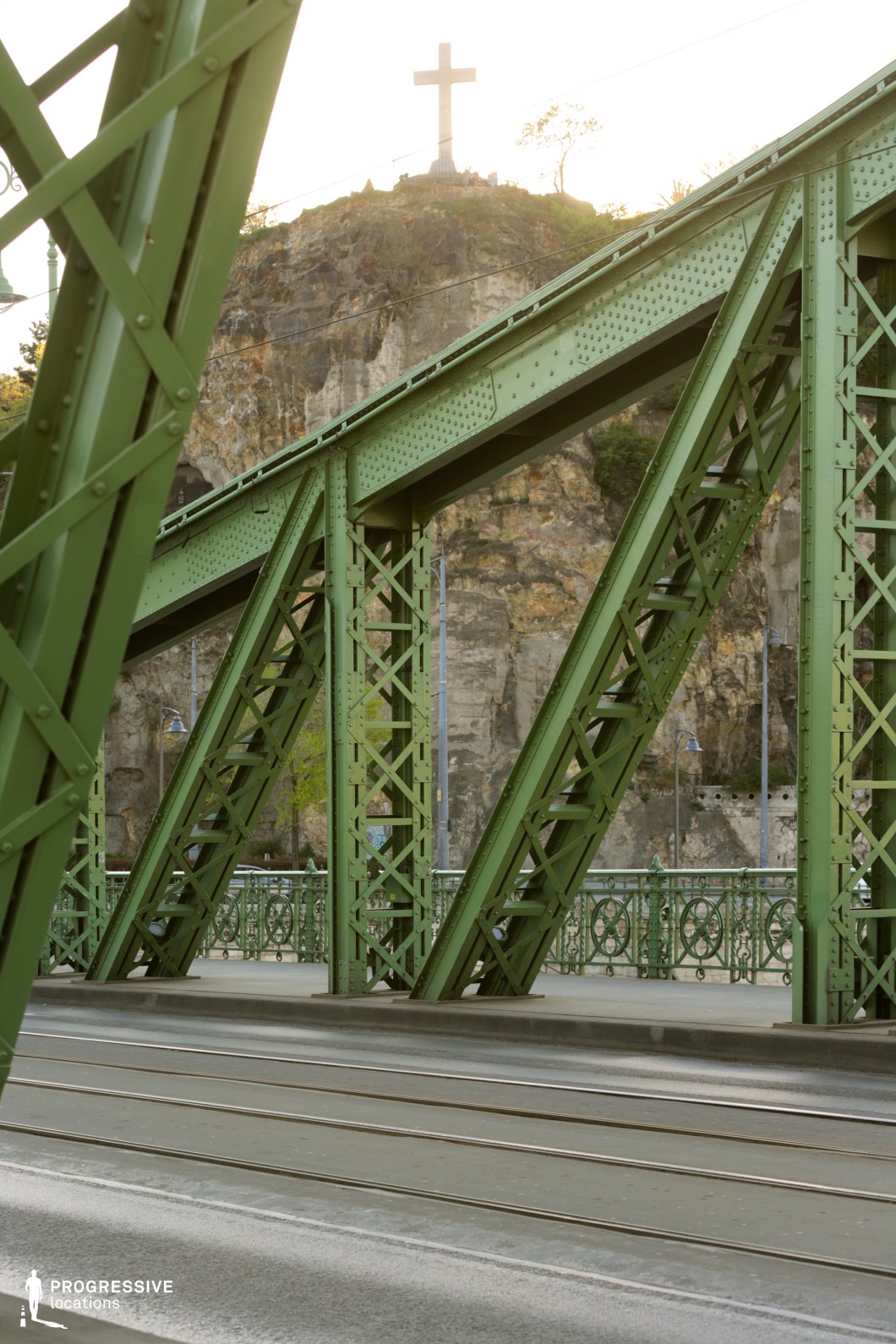 The image features a robust green iron bridge with intricate steel trusses, displaying the strength and architectural detail typical of industrial era structures. The bridge's design includes ornate railings and cross-bracing that highlight its functional yet aesthetic qualities. In the background, a prominent cross atop a rocky hill stands as a striking visual element against the natural rock face. This setting, combining industrial and natural elements, offers a dramatic backdrop for filming action sequences, historical or religious dramas, or urban thrillers, providing a location rich in symbolism and visual contrast.