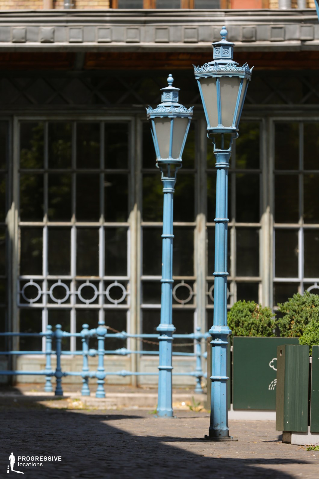 Two elegant blue wrought iron street lamps stand tall in the foreground, their intricate design adding charm to the setting. The lamps' slender poles are topped with ornate, vintage-style lanterns, complementing the surrounding blue railing that lines the cobblestone pathway. Behind them, large windows with detailed ironwork and an aged wooden frame create a historic, industrial atmosphere. Potted plants and greenery further enhance the peaceful yet timeless environment. This location, with its blend of vintage lighting and architectural elements, is perfect for filming scenes that require a serene, historical ambiance, ideal for dramas, period pieces, or romantic storylines.