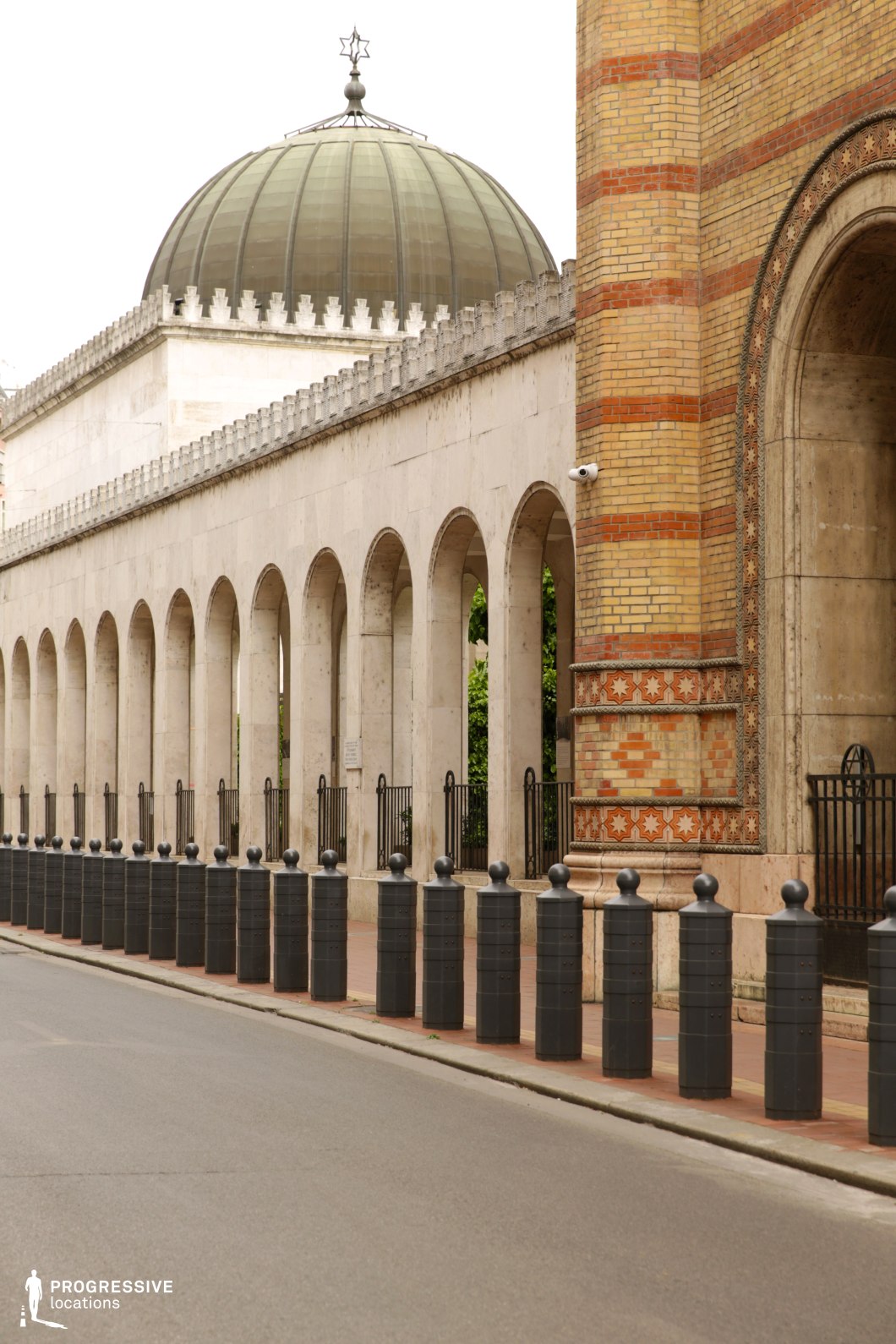 A grand, arching colonnade stretches along the street, composed of symmetrical stone arches, leading to a detailed brick column adorned with intricate star-shaped patterns. The domed structure in the background, topped with a decorative star, adds a striking focal point, complementing the architectural harmony of the space. The polished stone pavement is lined with cylindrical bollards, contributing to the orderly urban design. The juxtaposition of the bold, geometric lines of the arches and the rich, ornamental brickwork creates a timeless, classical atmosphere, ideal for capturing historical or dramatic scenes in films, particularly in urban or religious-themed narratives. This location offers versatility for films requiring grand architectural backdrops with an air of sophistication and significance.