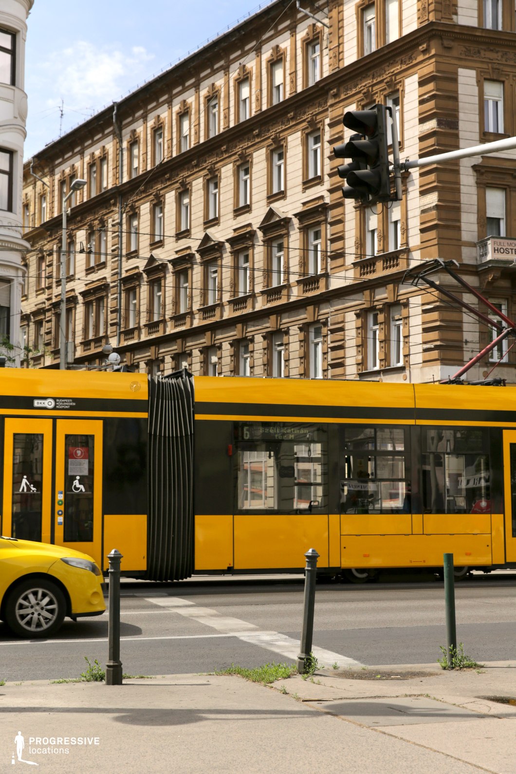 A modern yellow tram moves along an urban street, framed by historic, neoclassical buildings with detailed facades and symmetrical window arrangements. The tram's sleek, contemporary design contrasts with the ornate stonework of the background architecture, highlighting the blend of old and new in the cityscape. Overhead wires and a traffic light add to the dynamic urban environment, emphasizing the street’s function as a transit hub. This location is ideal for filming contemporary city life, fast-paced urban sequences, or stories that showcase the intersection of modern infrastructure and historical settings, making it suitable for dramas, thrillers, and documentary-style cinematography.