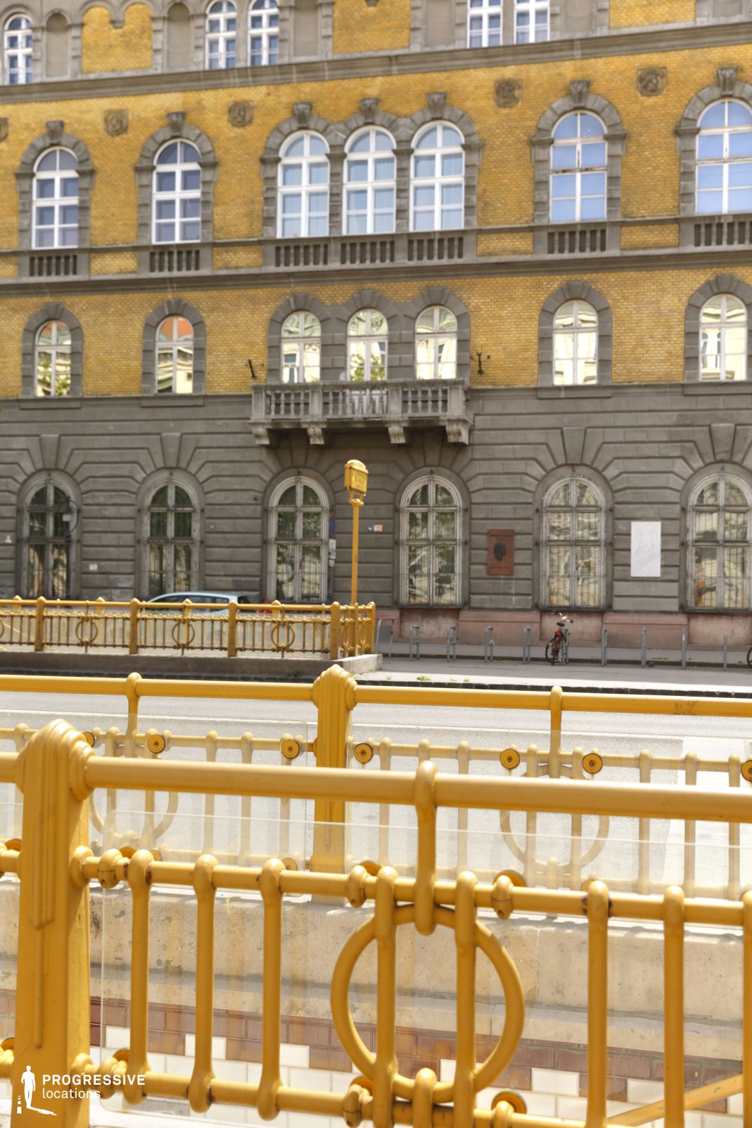 The image showcases a grand historic building with a striking yellow brick facade complemented by dark gray stone detailing. The symmetrical design features arched windows framed in stone, with decorative elements that enhance its classical architectural style. A small balcony with intricate stone railings is centrally positioned, adding to the building’s elegant aesthetic. In the foreground, a vibrant yellow metal railing with circular motifs runs along the edge of what appears to be a subway entrance or pedestrian underpass. The layered composition of railings, street elements, and the imposing facade creates a dynamic urban setting. This location is ideal for filming period dramas, historical narratives, or sophisticated cityscape shots. The depth and architectural details provide a visually rich backdrop for scenes involving urban exploration, mystery, or upscale metropolitan life.